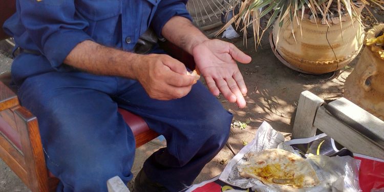 Long hours, low wages, and little dignity — a security guard pauses on duty to eat a simple, makeshift meal of roti with curry and a bowl of watery yogurt, placed on a worn bench in the open. With no proper dining space, no rest breaks, and minimal facilities, this scene reflects the daily reality of many guards in Pakistan who work exhausting shifts yet struggle to afford even a basic, nutritious meal.